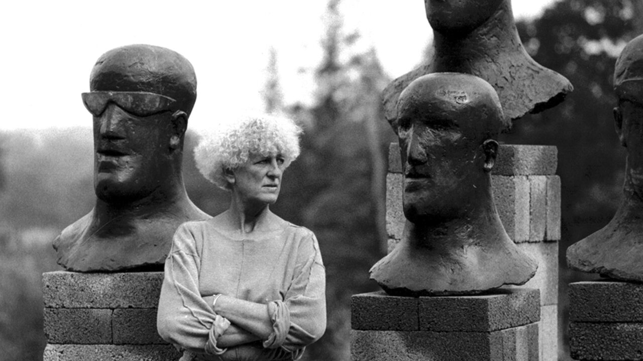 Elisabeth Frink standing with Goggle and Tribute heads, Woolland House grounds, 1984. Image © Christine Fauto/ Artist © in Elisabeth Frink images courtesy of Bree and Tully Jammet/ Courtesy of Dorset History Centre.